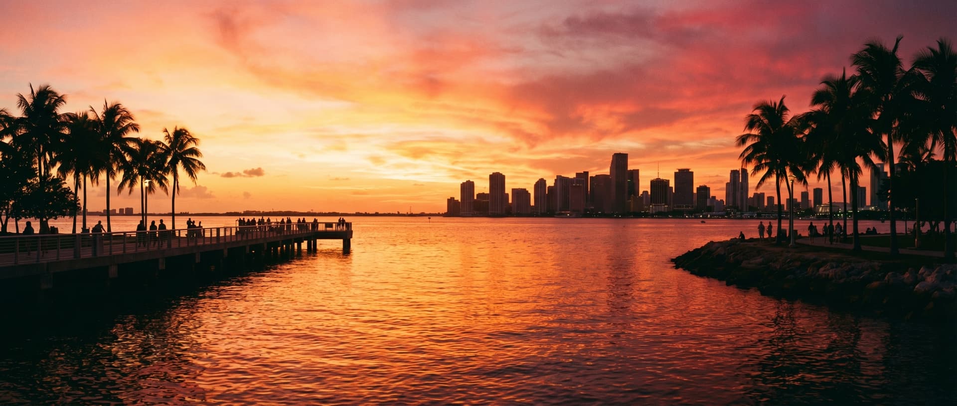 Panoramic view of Biscayne Bay at sunset from South Pointe Park pier in Miami Beach, golden orange and pink sky reflecting on calm bay water, silhouetted palm trees framing the shot, downtown Miami skyline in the distance