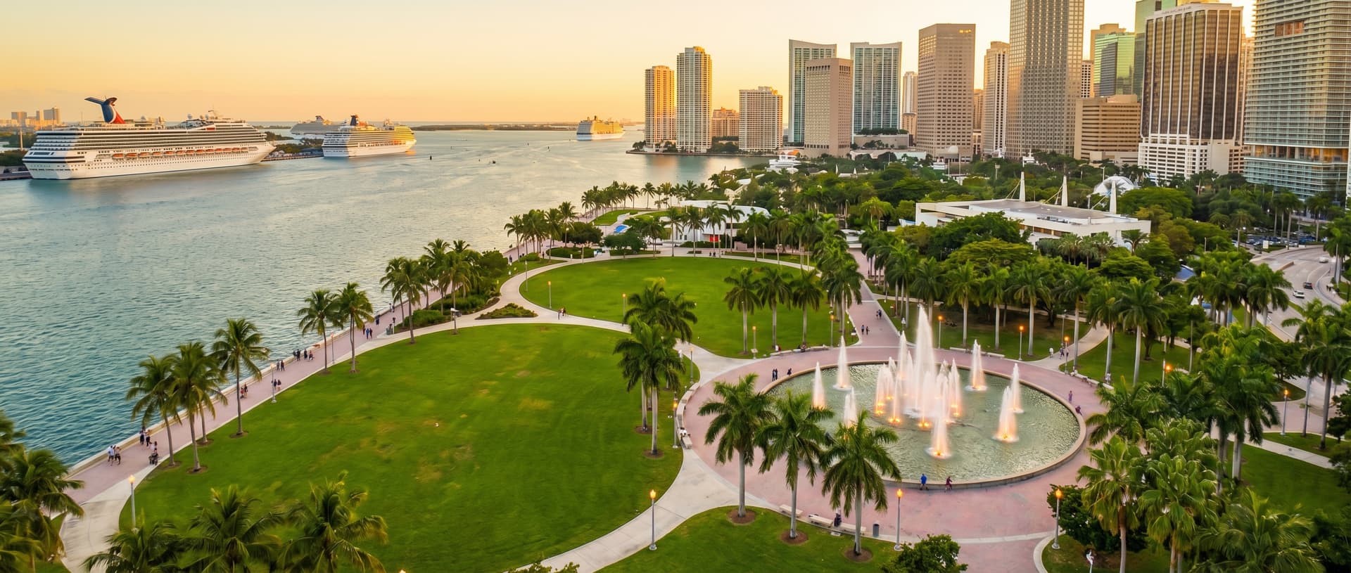 Aerial view of Bayfront Park in downtown Miami at golden hour, a 32-acre green waterfront park along Biscayne Bay with winding walkways, the Pepper Fountain, palm trees and people on the baywalk, with downtown Miami skyscrapers and cruise ships in the background