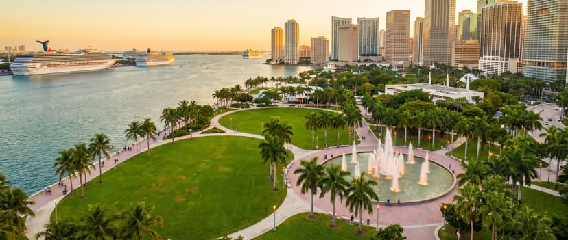 Aerial view of Bayfront Park in downtown Miami at golden hour, a 32-acre green waterfront park along Biscayne Bay with winding walkways, the Pepper Fountain, palm trees and people on the baywalk, with downtown Miami skyscrapers and cruise ships in the background