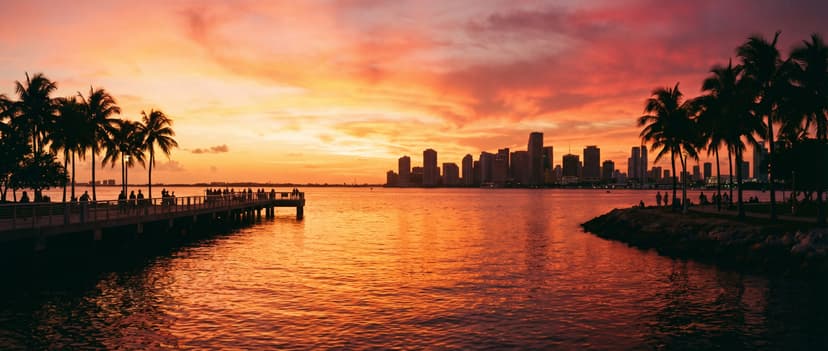 Panoramic view of Biscayne Bay at sunset from South Pointe Park pier in Miami Beach, golden orange and pink sky reflecting on calm bay water, silhouetted palm trees framing the shot, downtown Miami skyline in the distance