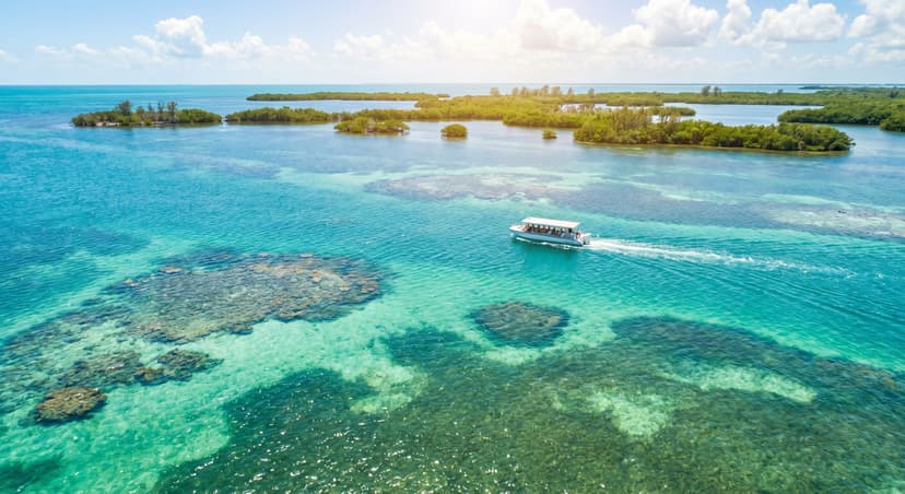 Aerial view of turquoise Biscayne Bay with coral reefs visible through crystal clear shallow water, mangrove islands in the background, and a small tour boat on the water under tropical Florida sunshine