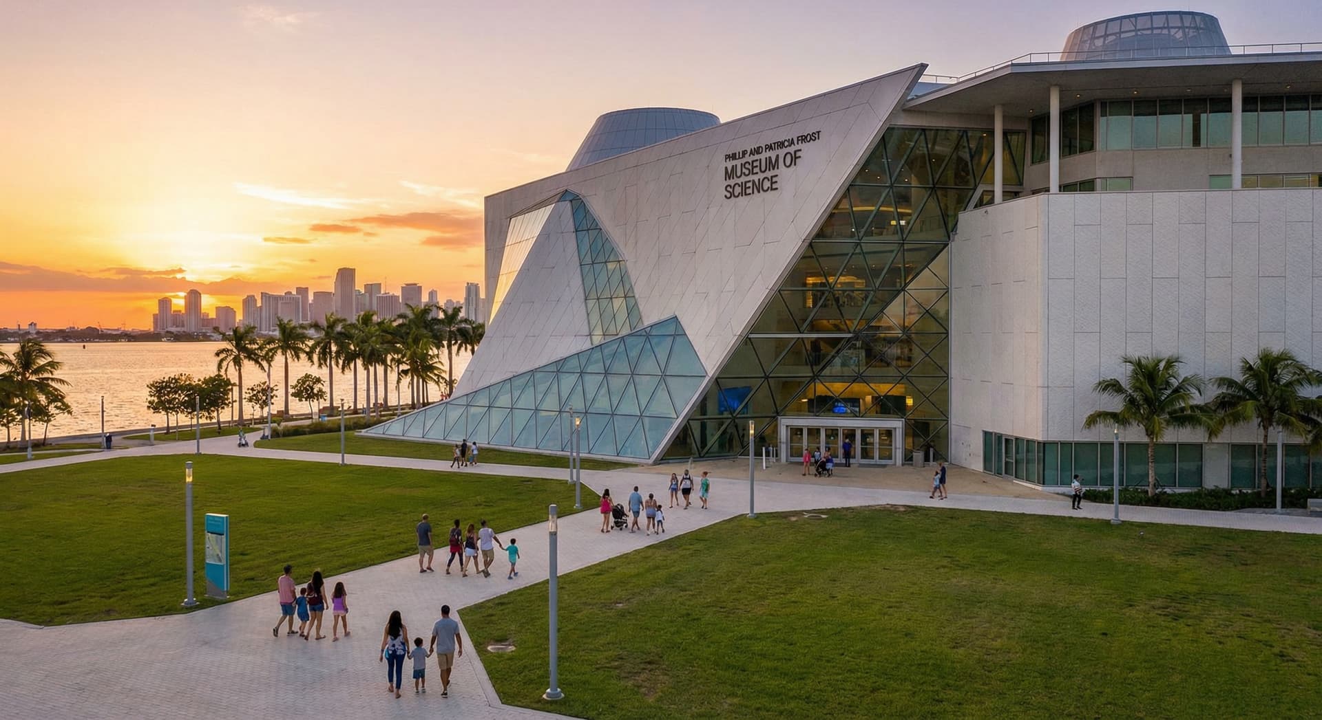 Exterior of the Phillip and Patricia Frost Museum of Science in Miami at golden hour with modern angular white architecture, geometric glass panels, Museum Park green lawn, Biscayne Bay sparkling in the background, downtown Miami skyline, and palm trees