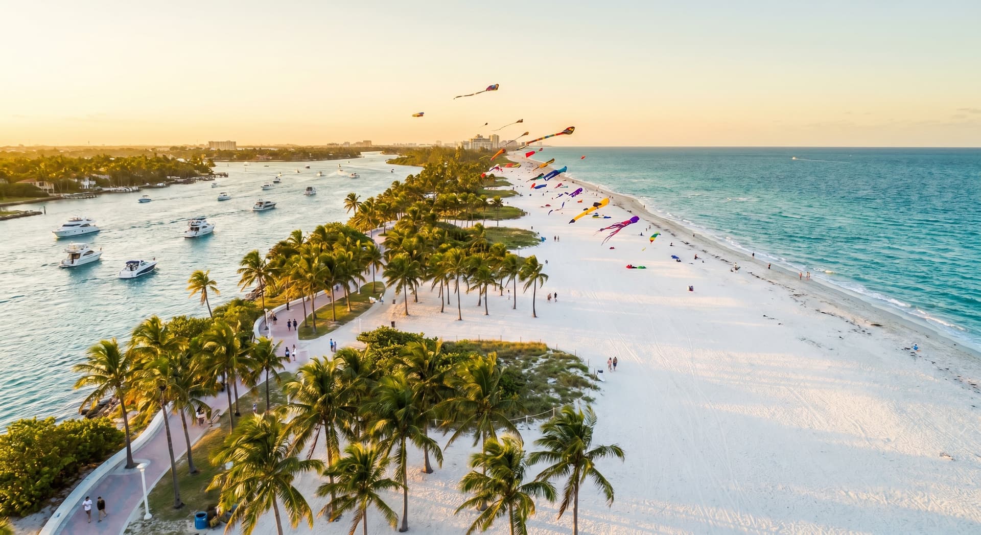 Aerial view of Haulover Beach Park in Miami at golden hour with pristine white sand meeting turquoise Atlantic waters, colorful kites flying above, palm trees lining the park, and boats near the inlet