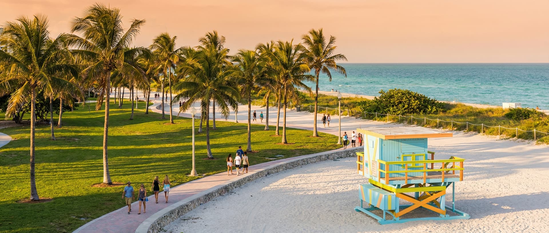 Wide view of Lummus Park in South Beach Miami at golden hour with lush green grass, tall coconut palm trees, a colorful Art Deco lifeguard tower, white sand beach, and turquoise Atlantic Ocean in the background