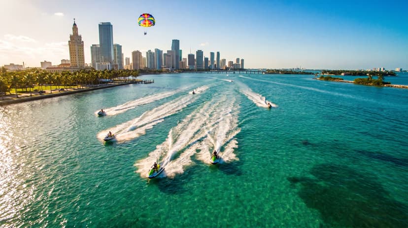 Aerial view of jet skiers riding across turquoise Biscayne Bay in Miami with the downtown skyline in the background and a parasail visible in the golden hour sky