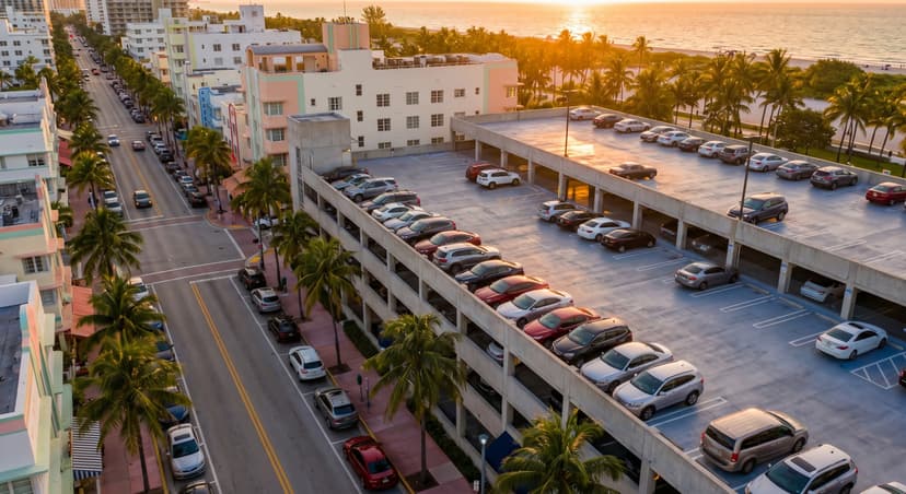 Aerial view of a Miami Beach parking garage and Collins Avenue at golden hour, with rows of parked cars, art deco buildings, and palm trees lining the tropical urban street